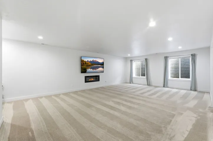 Spacious room featuring light-toned carpeting with subtle stripes, recessed lighting, and a wall-mounted linear fireplace