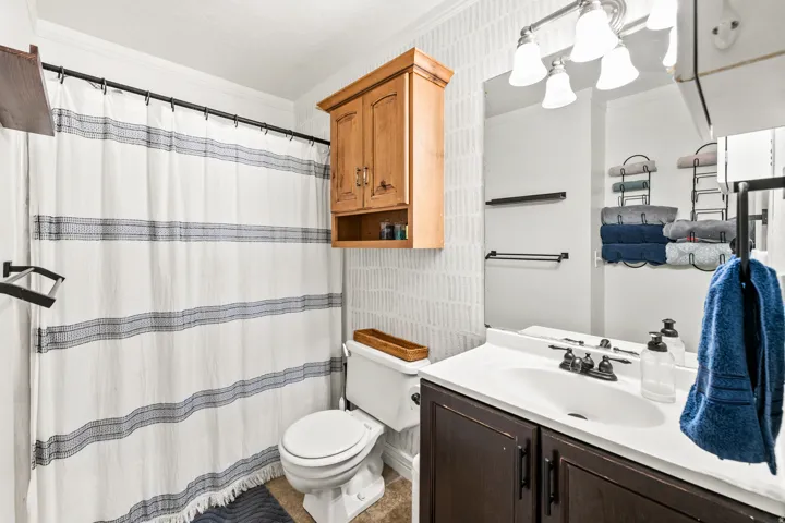 Bathroom featuring a dark wood-finish vanity with an integrated white sink, a vanity mirror with an overhead three-bulb light fixture, a toilet, and an over-toilet wood-finish cabinet
