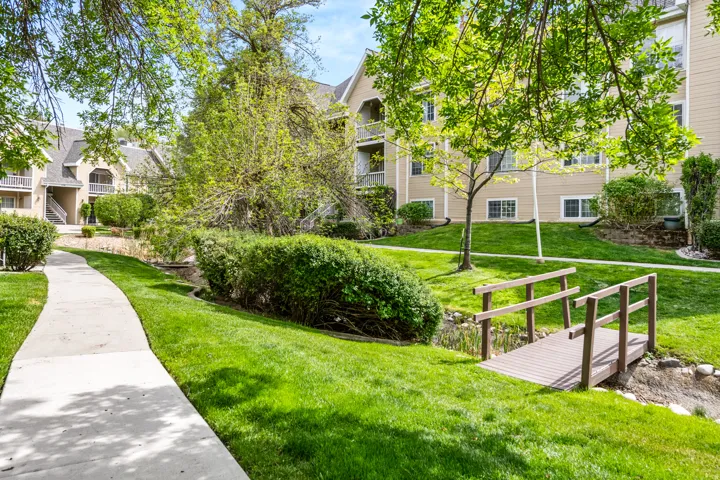 Manicured green space featuring a concrete pathway, a decorative wooden bridge over a dry creek bed, mature trees, and shrubbery