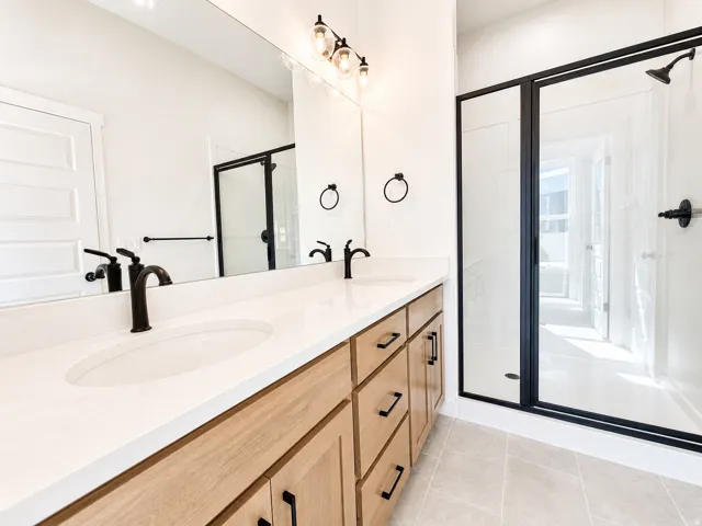 Contemporary bathroom featuring a dual vanity with light wood-finish cabinetry, white countertops, and matte black fixtures