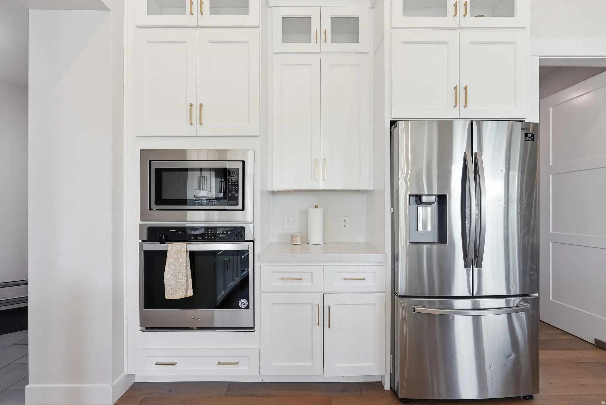 Kitchen featuring white shaker-style cabinetry with gold-finish hardware, built-in stainless steel microwave and wall oven, stainless steel French door refrigerator with ice and water dispenser, and light-toned countertops