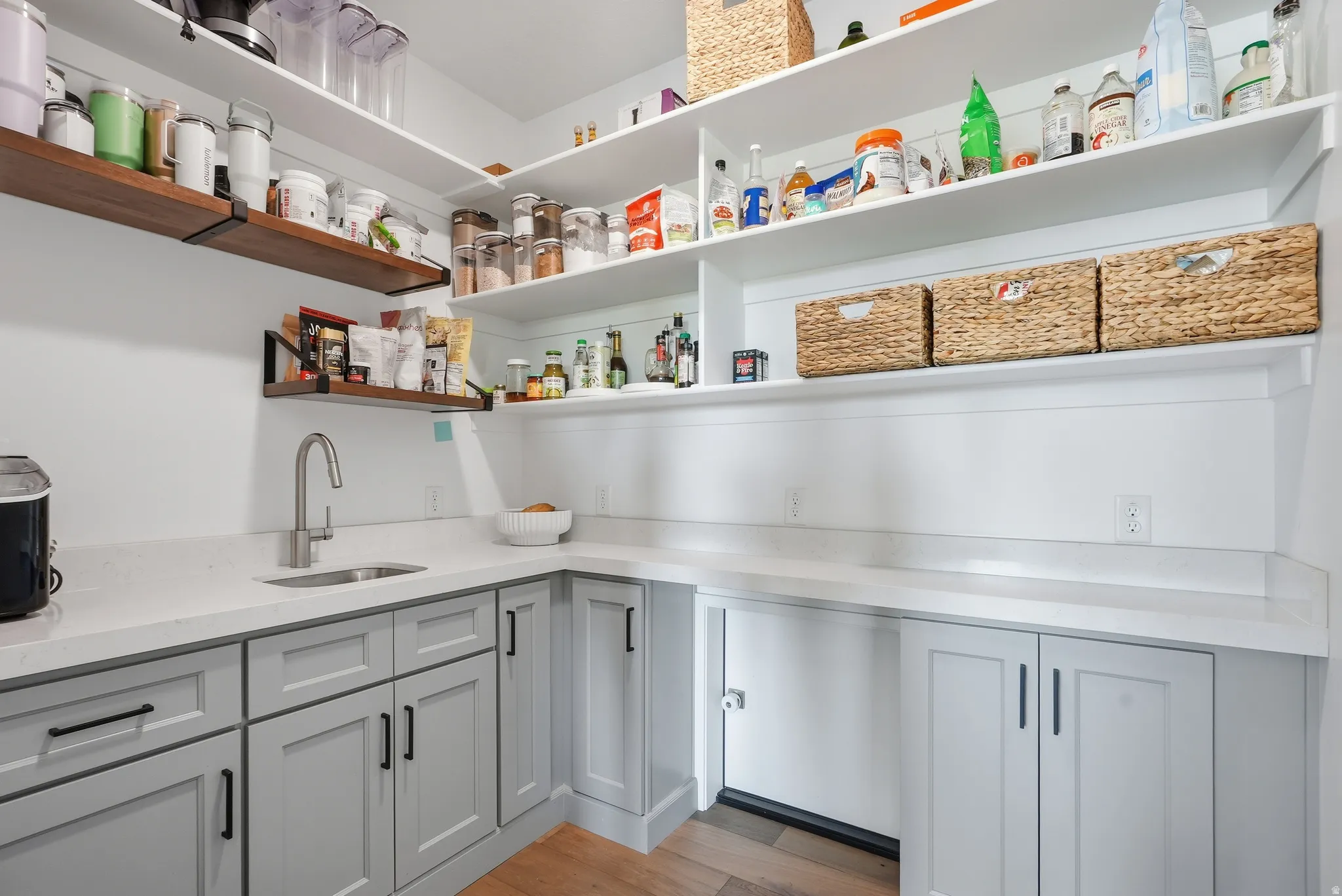 Spacious pantry featuring extensive white shelving, light gray cabinetry, a built-in sink with a gooseneck faucet, light-toned countertops, and wood-finish flooring