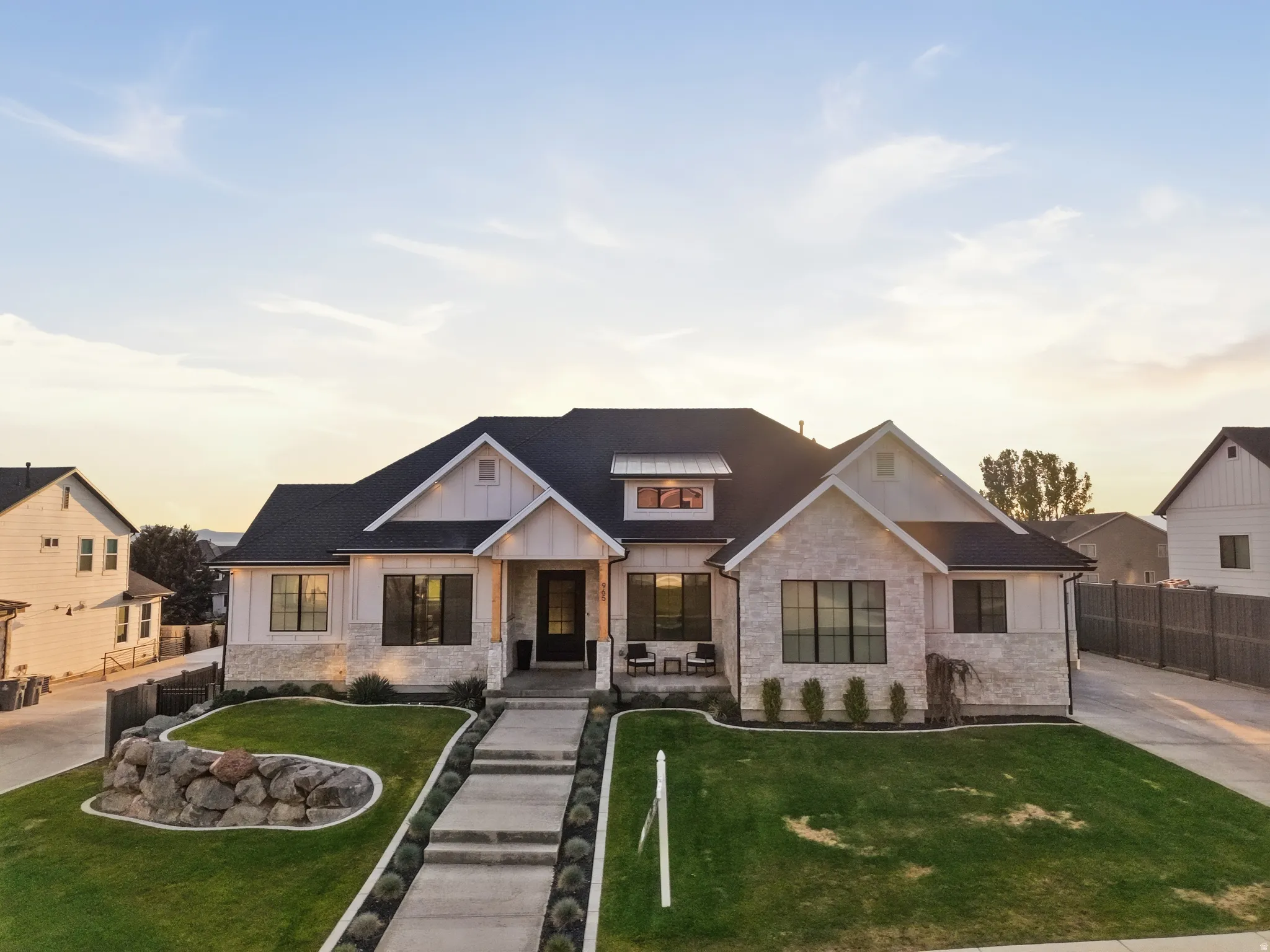 Modern farmhouse exterior featuring board and batten siding, stone accents, dark-framed windows, a black asphalt shingle roof, and a concrete walkway bordered by manicured landscaping