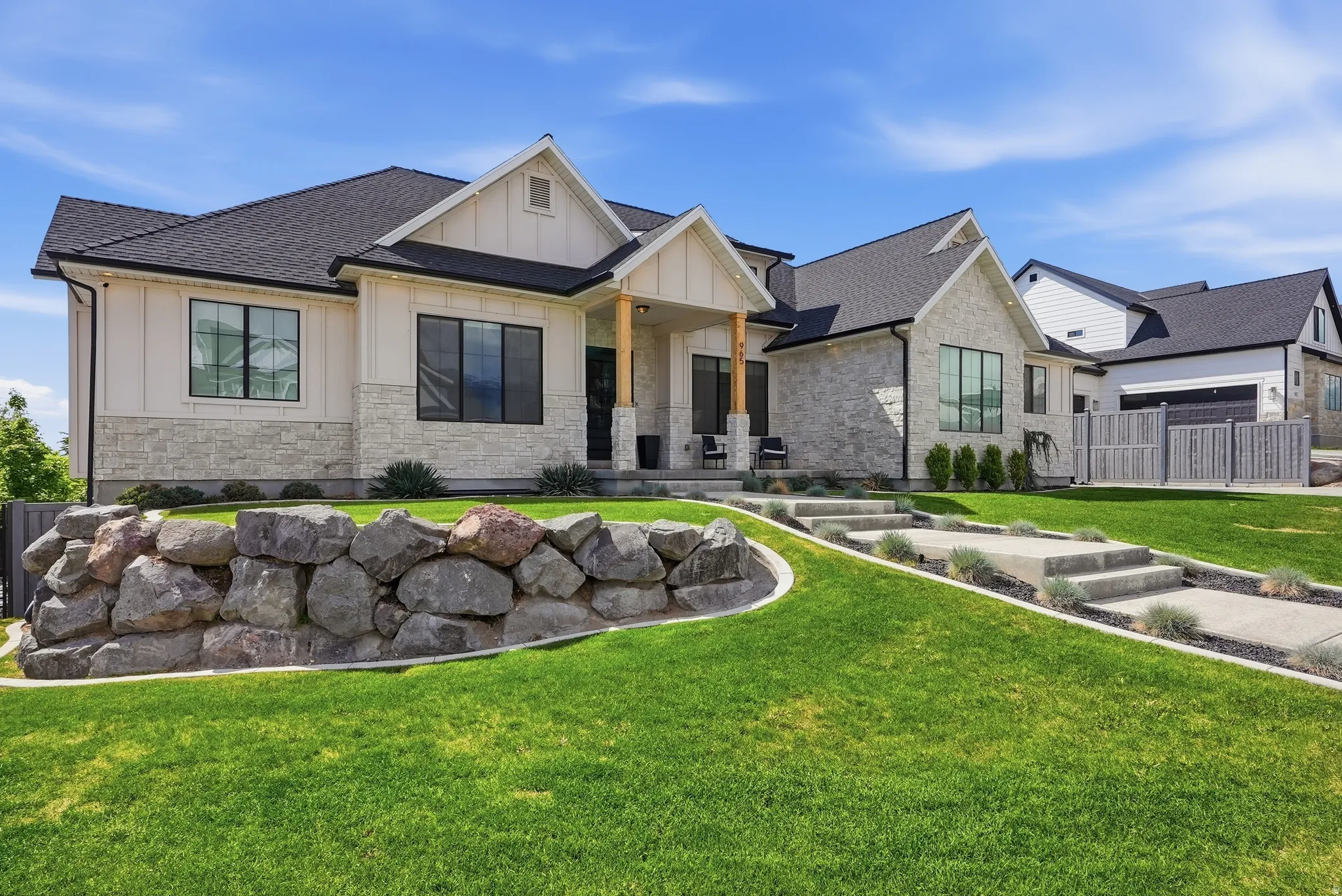 Modern farmhouse exterior featuring board and batten siding, stone accents, black-framed windows, a covered entry with wood-wrapped columns, and a terraced front yard with a rock retaining wall