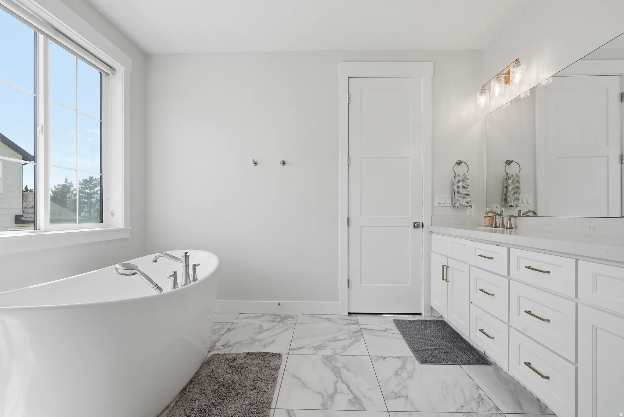 Freestanding soaking tub with floor-mounted fixtures, large window, white vanity with dual sinks, quartz countertop, and large mirror with sconce lighting