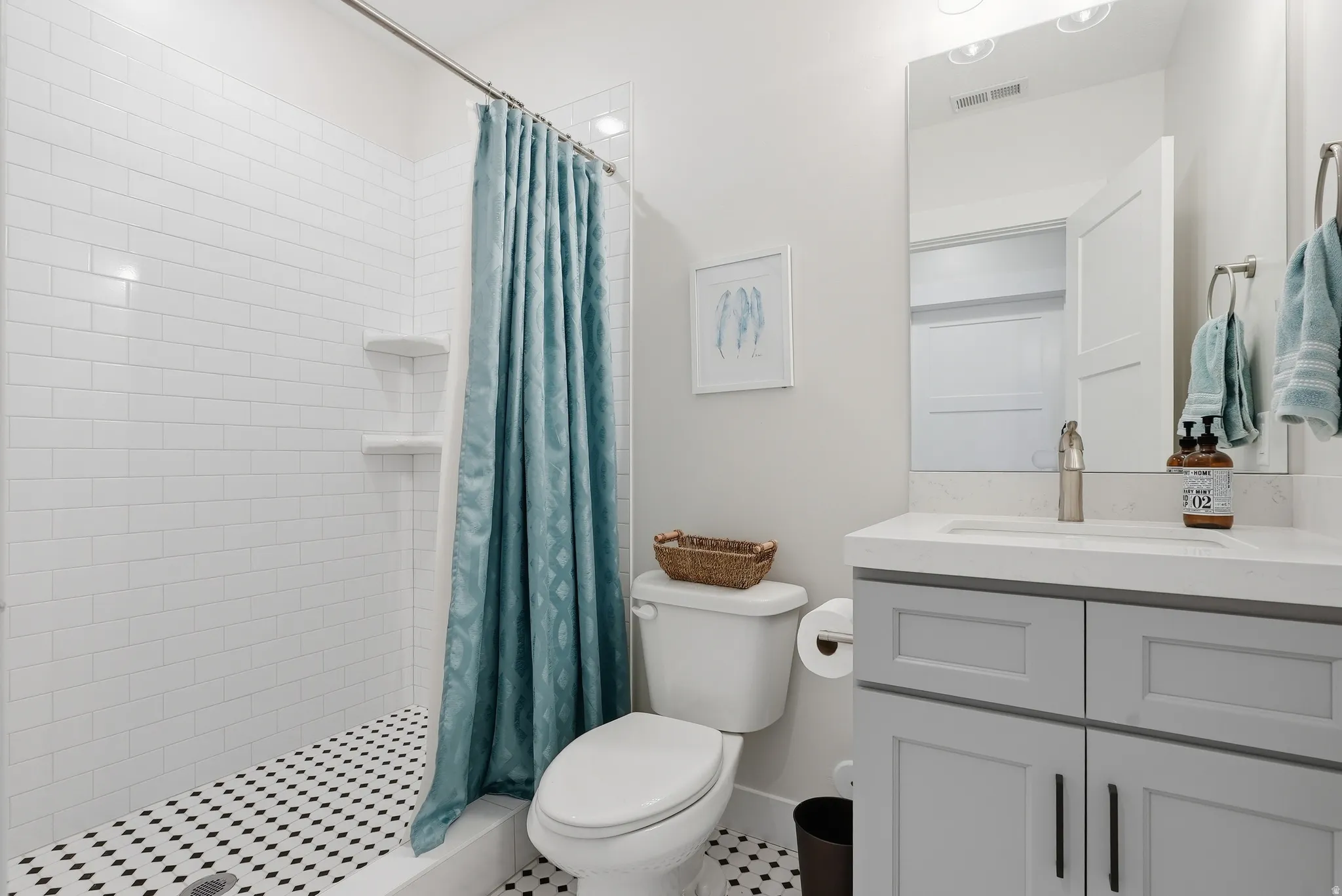 Bathroom featuring a subway-tiled shower with black and white mosaic flooring, a white toilet, and a vanity with a white countertop, undermount sink, and light gray cabinetry