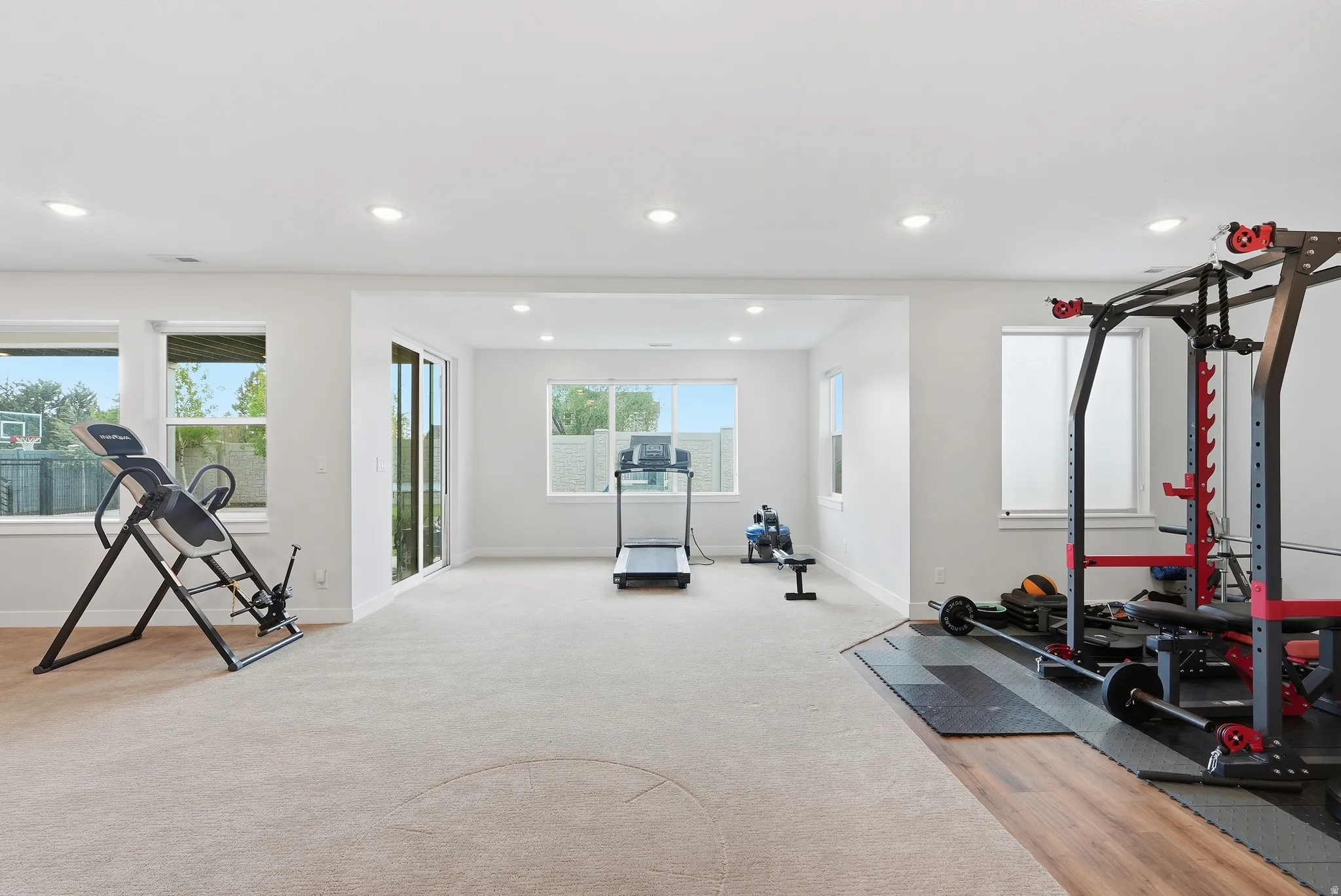 Spacious room featuring light-toned carpeting and wood-finish flooring sections, multiple large windows, recessed ceiling lighting, and a sliding glass door