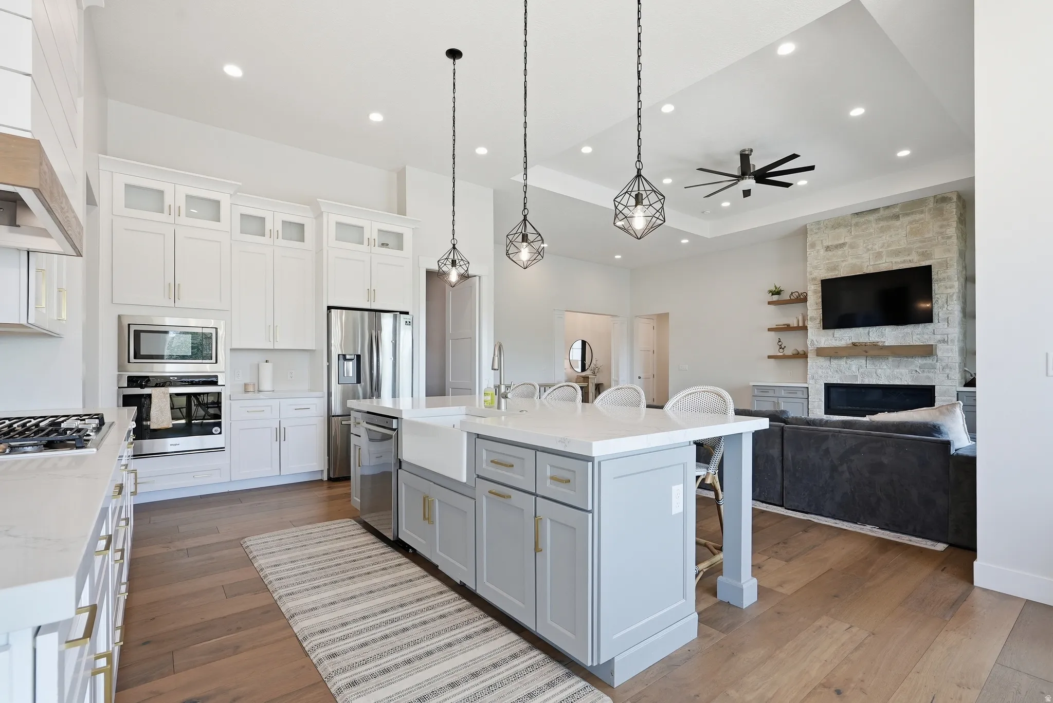 Open-concept kitchen and living area featuring wood-finish flooring, a kitchen island with seating, pendant lighting, stainless steel appliances, and a stone fireplace