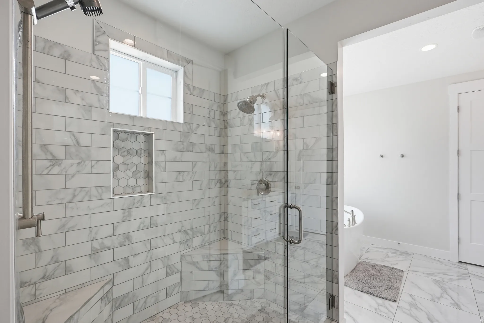 Spacious walk-in shower featuring a glass enclosure, built-in shelving with hexagonal tile accent, a fixed window, and a corner bench
