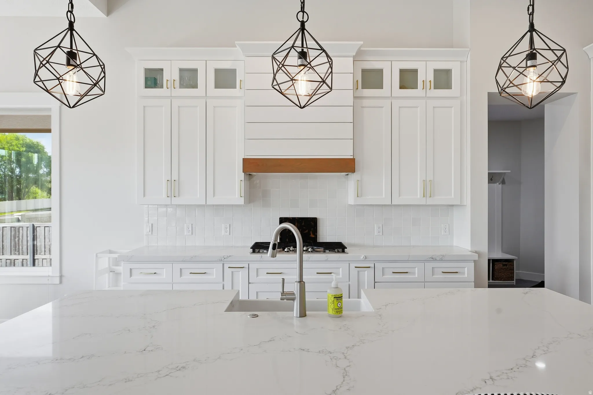 Spacious kitchen featuring an expansive island with an undermount sink, white cabinetry with upper glass-front accents, marble-finish countertops, a subway tile backsplash, and decorative geometric pendant lighting