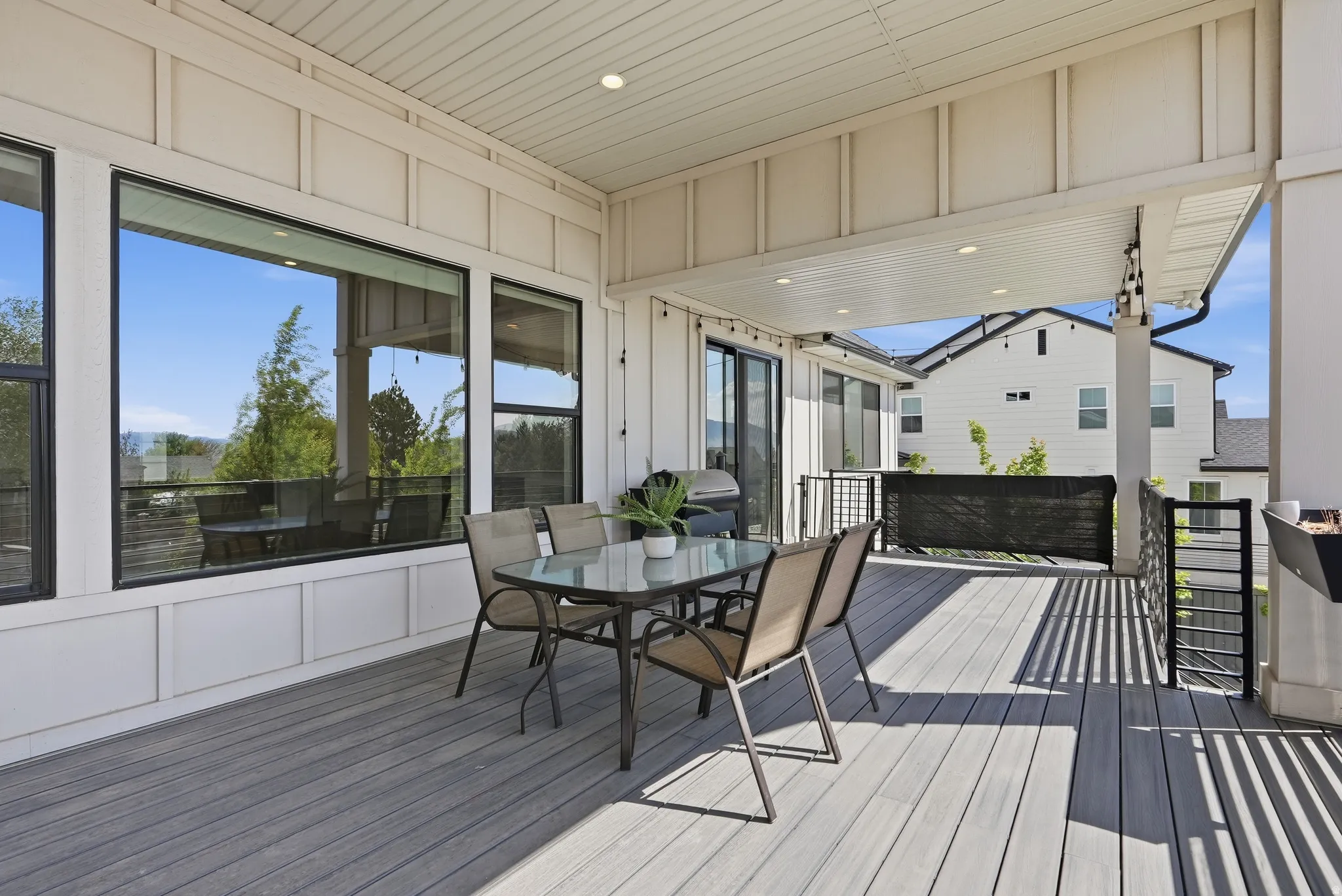 Covered outdoor deck featuring composite decking, recessed lighting, white shiplap siding, expansive windows, and a black metal railing