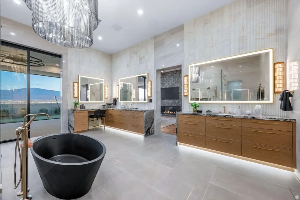 Spacious bathroom featuring a freestanding black soaking tub, dual floating wood-finish vanities with stone countertops, backlit mirrors, and a large multi-tiered chandelier
