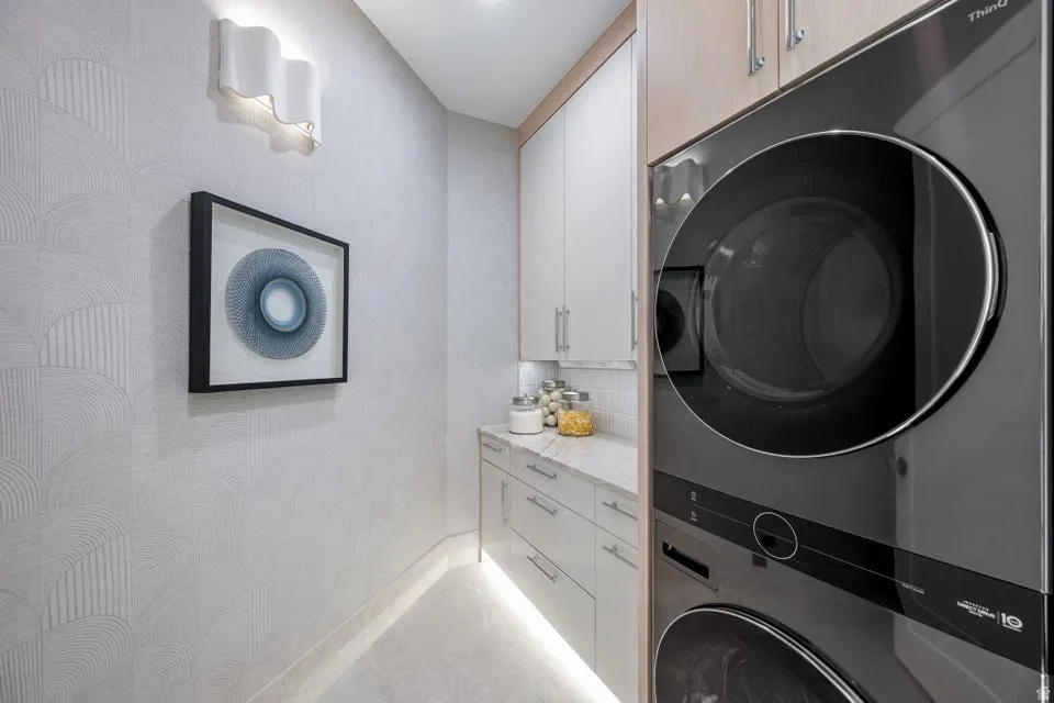 Integrated laundry area featuring a stacked washer and dryer, under-cabinet lighting, contemporary wall sconce, upper and lower cabinetry, and a light-toned countertop