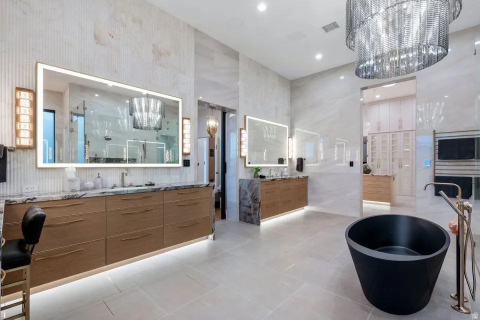 Spacious bathroom featuring a freestanding black soaking tub, dual wood-finish vanity cabinets with stone countertops, back-lit mirrors, and large format tile flooring