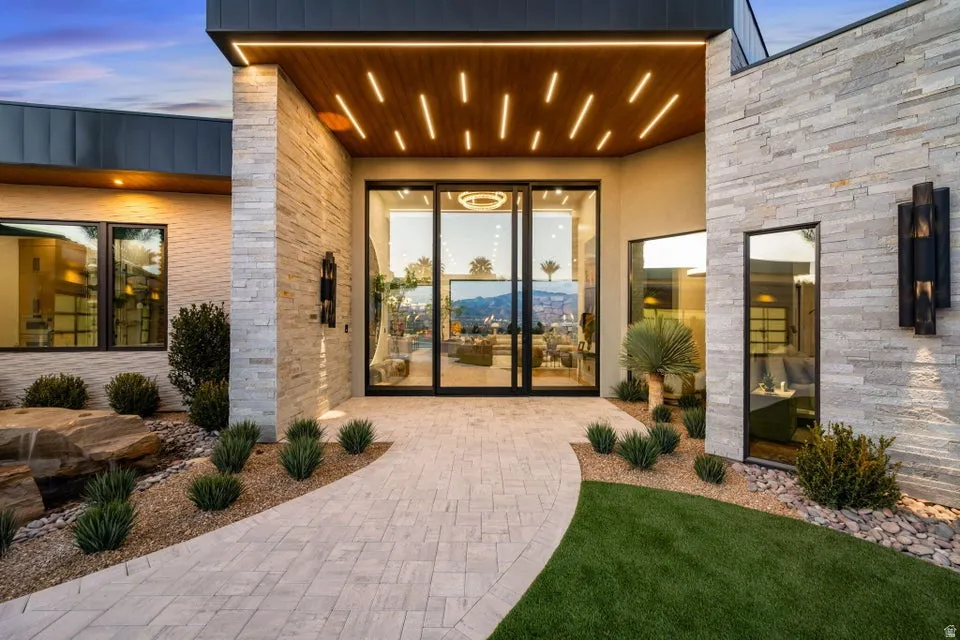 Modern exterior design featuring natural stone accents, a wood-paneled entry ceiling with recessed lighting, full-light entry doors, a paver pathway, and curated landscaping