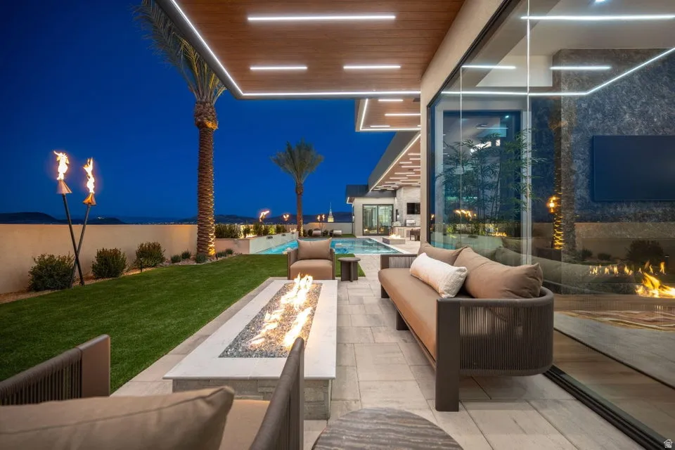 Expansive outdoor living area featuring a built-in linear fire pit, a covered patio with recessed lighting, and a manicured lawn