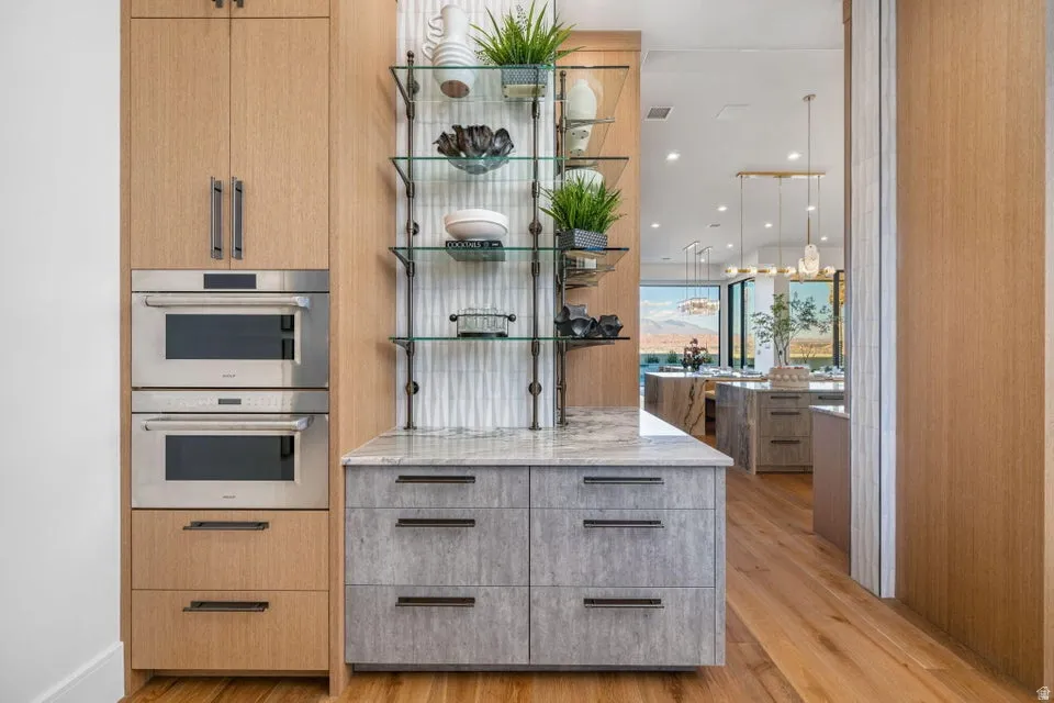Built-in stainless steel double ovens, custom wood-finish cabinetry, gray textured lower cabinetry with dark hardware, glass shelving with dark metal supports, and a light-toned countertop
