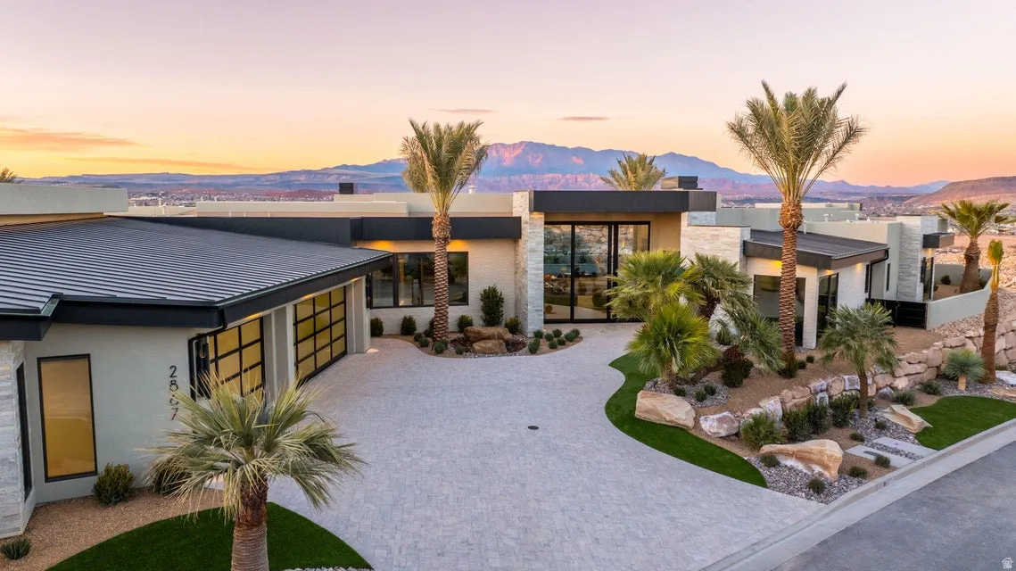 Contemporary desert-modern residence featuring a paver driveway, integrated landscaping with mature palm trees, a flat roofline, and expansive glass doors