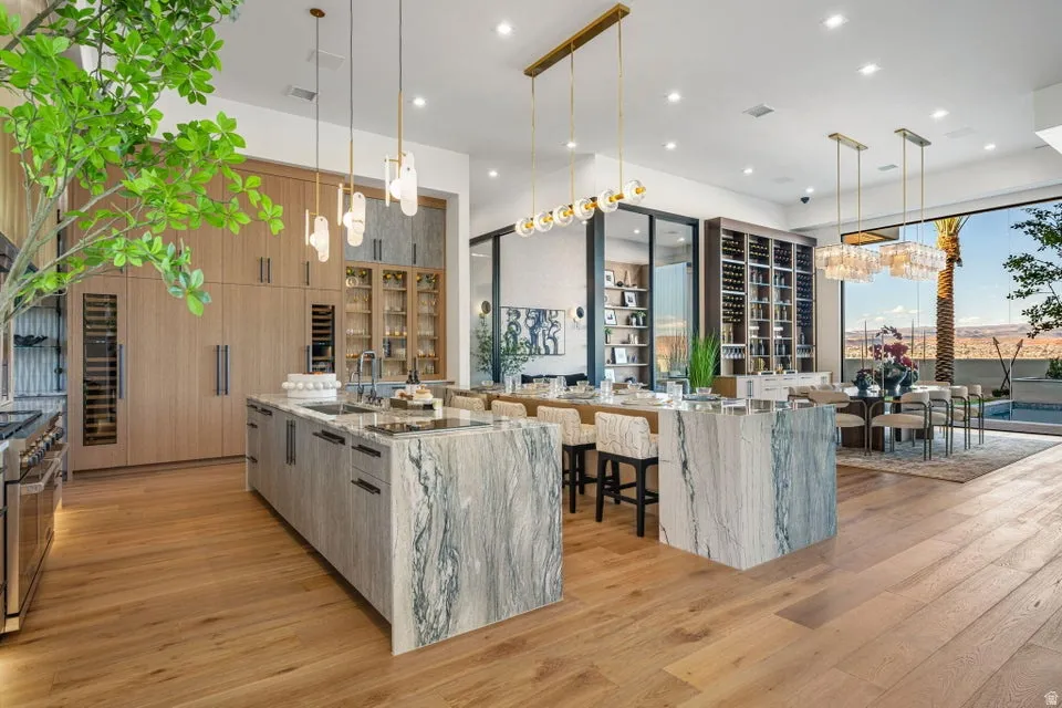 Open-concept kitchen and dining area featuring wood-finish flooring, two large stone islands, custom light wood cabinetry, and a full-height wine storage display