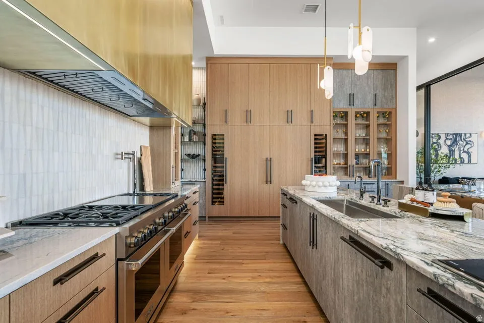 Gourmet kitchen featuring a commercial-grade range, brass hood, natural wood-finish cabinetry, stone countertops, and integrated wine refrigerators