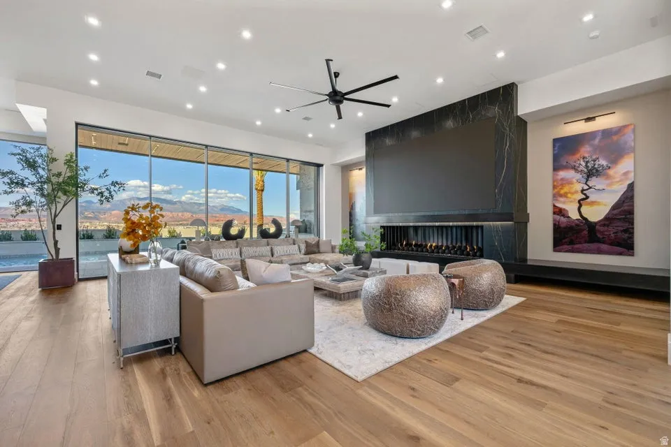 Living area with full-height glass walls showcasing mountain and desert scenery, a linear fireplace with a dark stone surround, wood-finish flooring, a contemporary ceiling fan, and recessed lighting