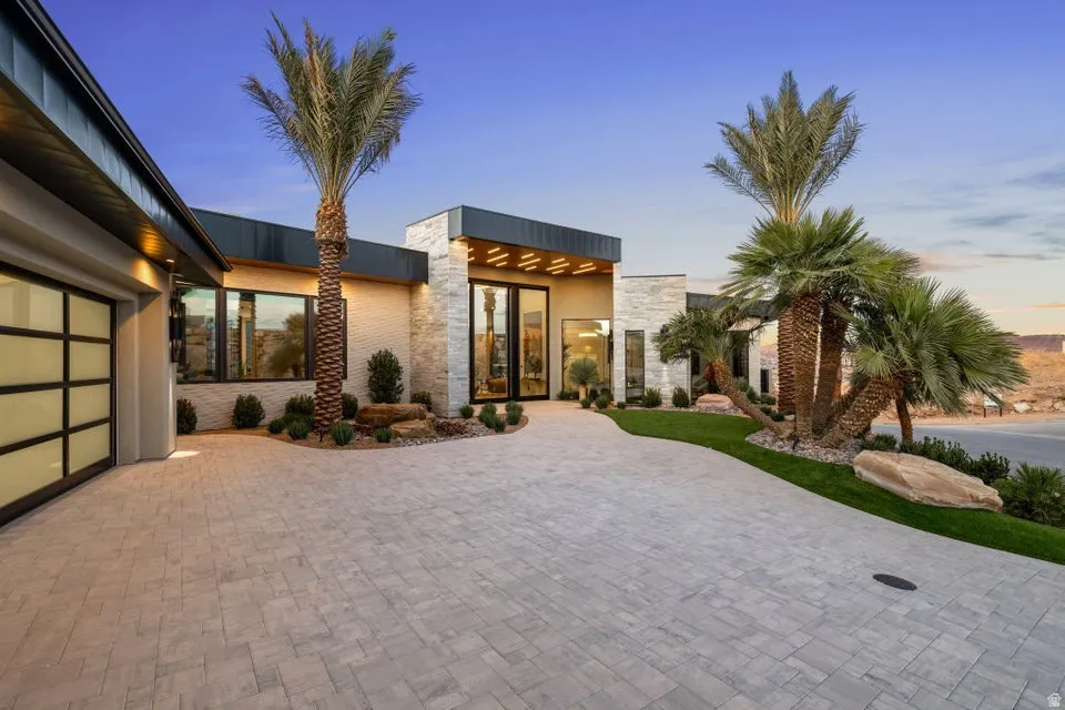 Contemporary exterior featuring a stone facade, a multi-pane glass garage door, a paver driveway, mature palm trees, and manicured landscaping