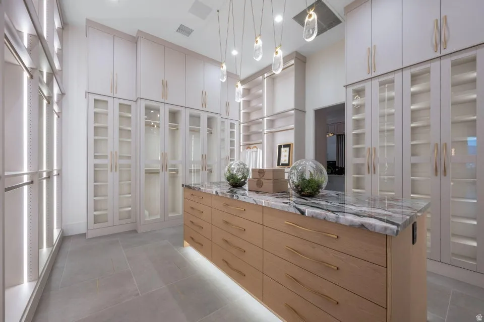 Spacious custom closet featuring extensive cabinetry with glass panel doors, integrated lighting, a large central island with a stone countertop and wood-finish drawers, recessed ceiling lights, and tile flooring