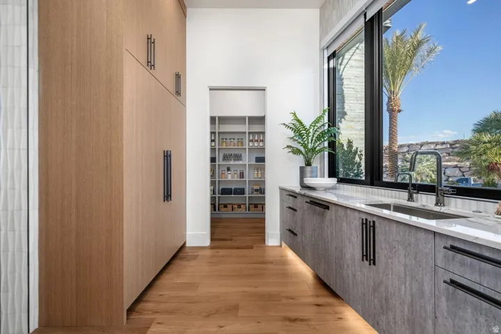 Modern kitchen featuring wood-finish flooring, full-height custom cabinetry, a built-in double sink, a large window, and a walk-in pantry with extensive shelving