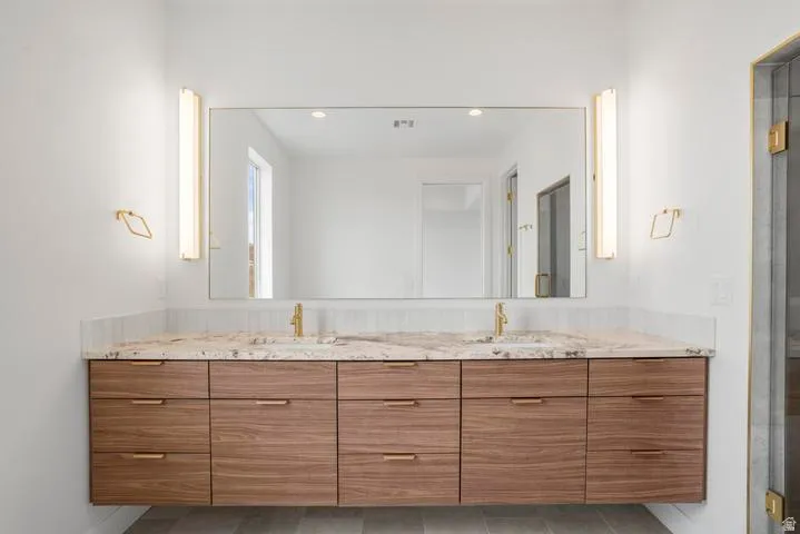 Dual basin vanity featuring a wood-finish floating cabinet with gold-tone hardware, a stone countertop, and a full-width mirror framed by vertical sconces