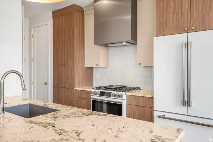 Kitchen featuring a deep undermount sink with a gooseneck faucet, light-toned stone countertops, a stainless steel range with a ventilation hood, and two-tone cabinetry with wood-finish and cream-colored doors