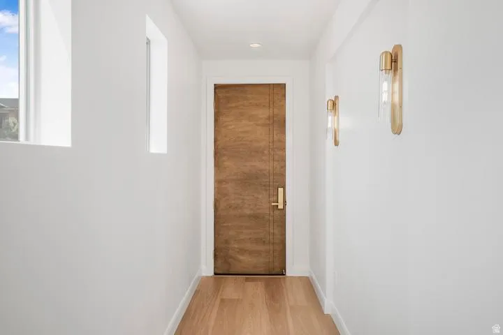 Hallway featuring a natural wood door with brass hardware, light wood-finish flooring, two rectangular windows, and modern wall sconces