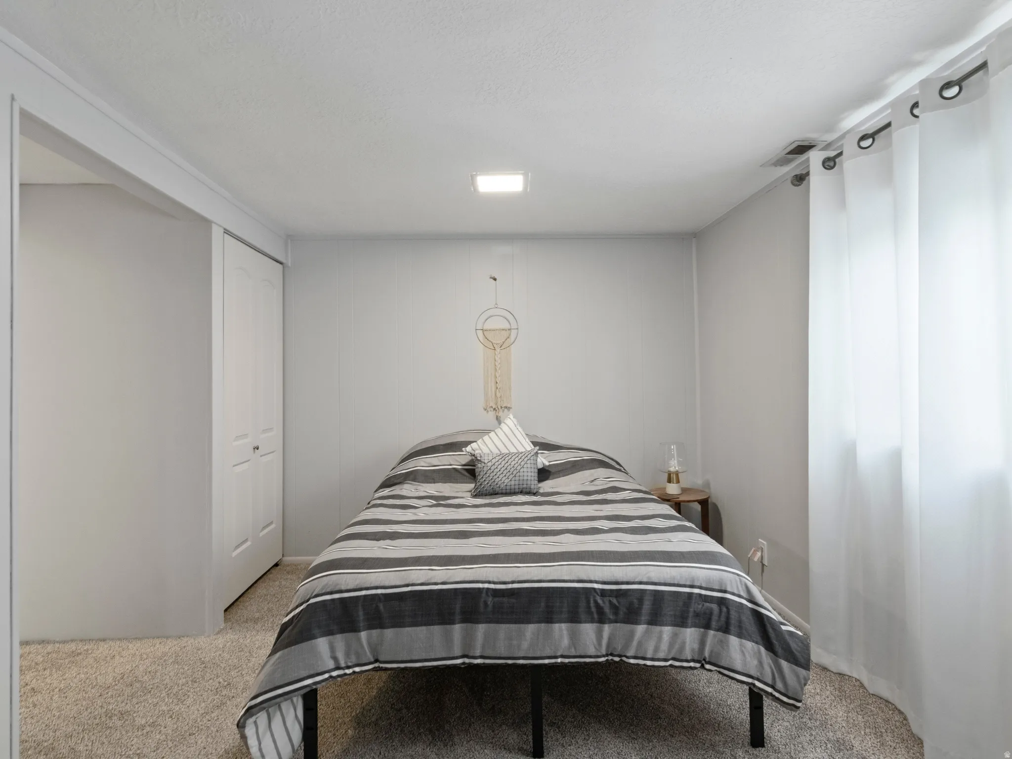 Studio bedroom with Carpeted floors. Featuring a white bi-fold closet door, textured ceiling, recessed lighting, and a large windows with white curtains
