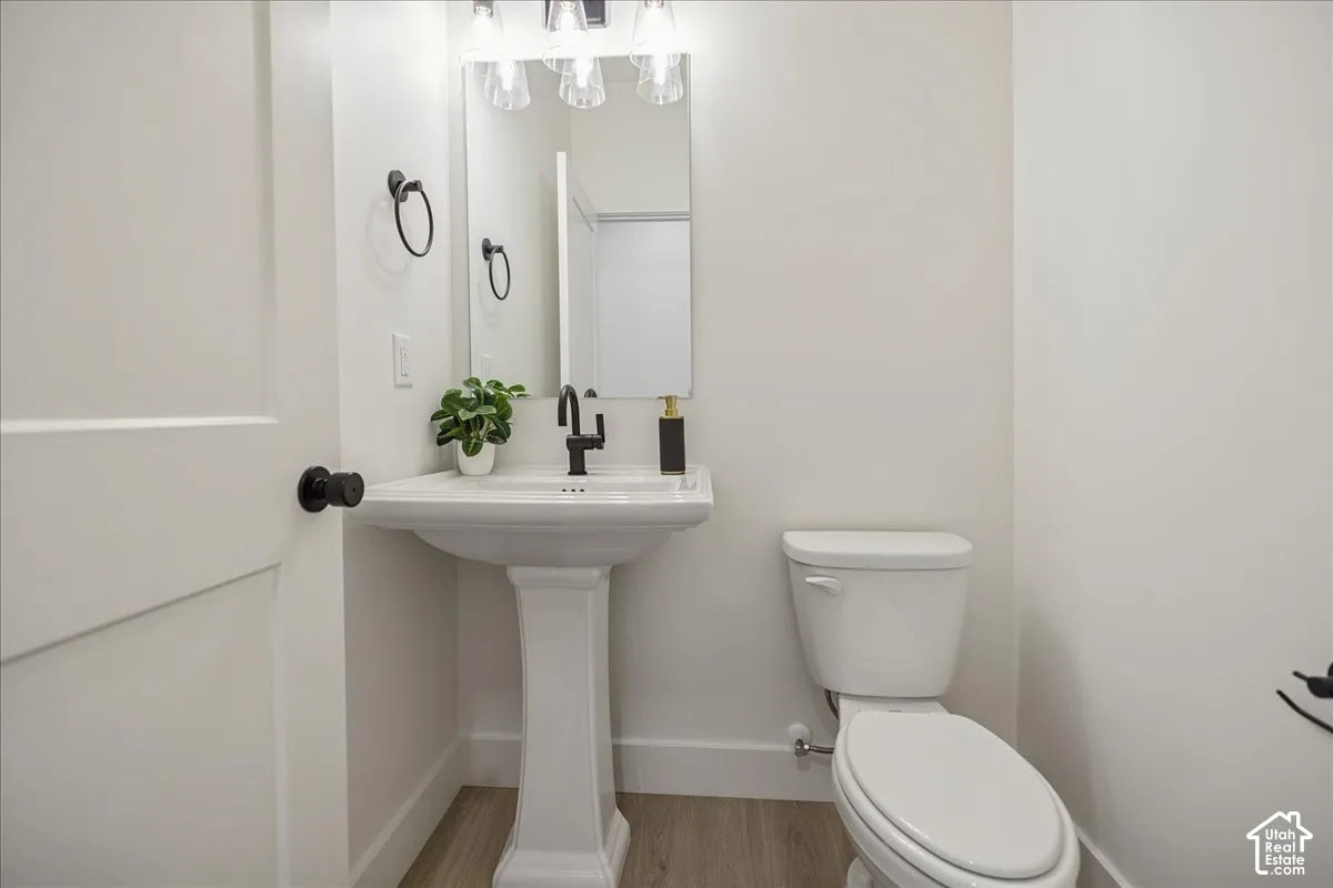 Pedestal sink with matte black faucet, wall-mounted mirror, three-light fixture, wood-finish flooring, and white toilet