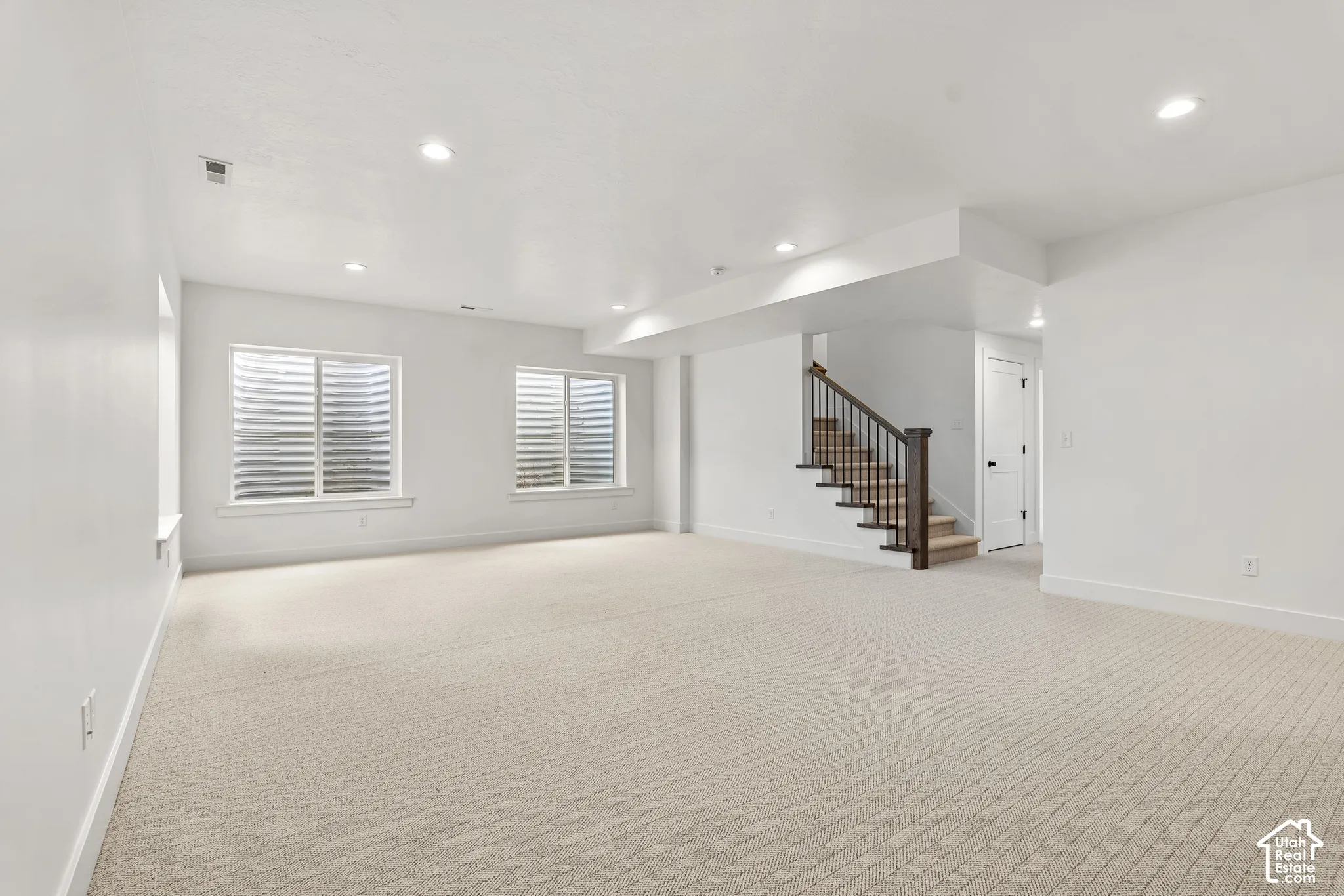 Spacious room featuring neutral carpet flooring, recessed lighting, two egress windows with horizontal blinds, a staircase with dark wood-finish newel posts and railings, and white trim