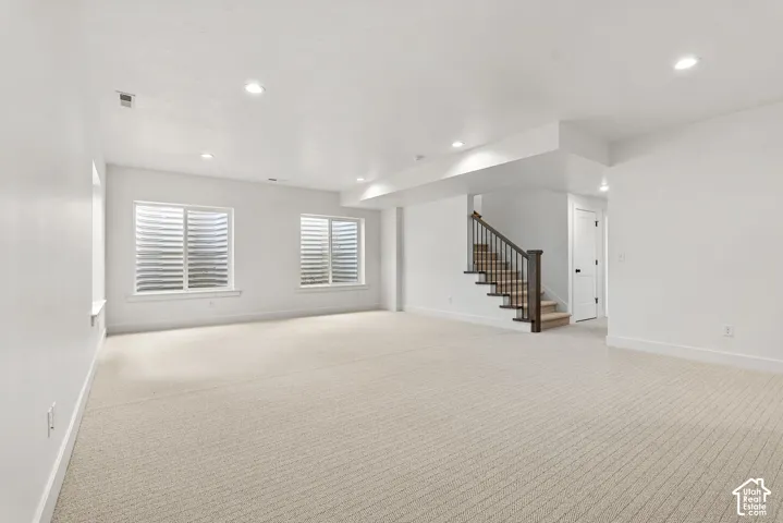 Spacious room featuring neutral carpet flooring, recessed lighting, two egress windows with horizontal blinds, a staircase with dark wood-finish newel posts and railings, and white trim