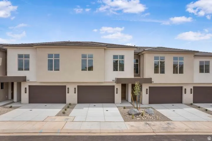 Modern stucco townhome exterior featuring an attached two-car garage, large windows, an integrated front entry awning, and a contemporary landscape design with decorative rock