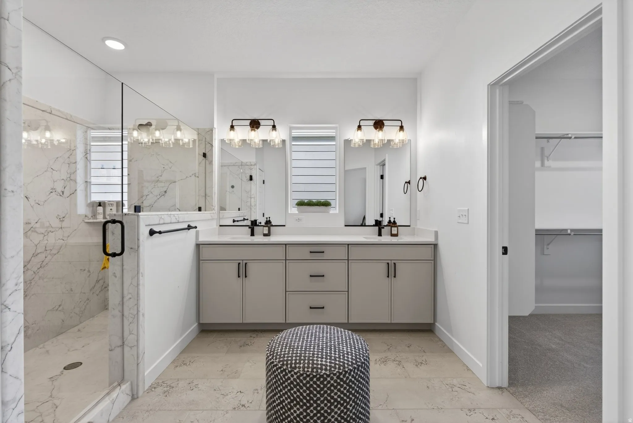 Contemporary bathroom featuring a spacious glass-enclosed shower with marble-look tile, a dual vanity with light gray cabinetry and white countertops, dual framed mirrors, and stylish wall-mounted light fixtures
