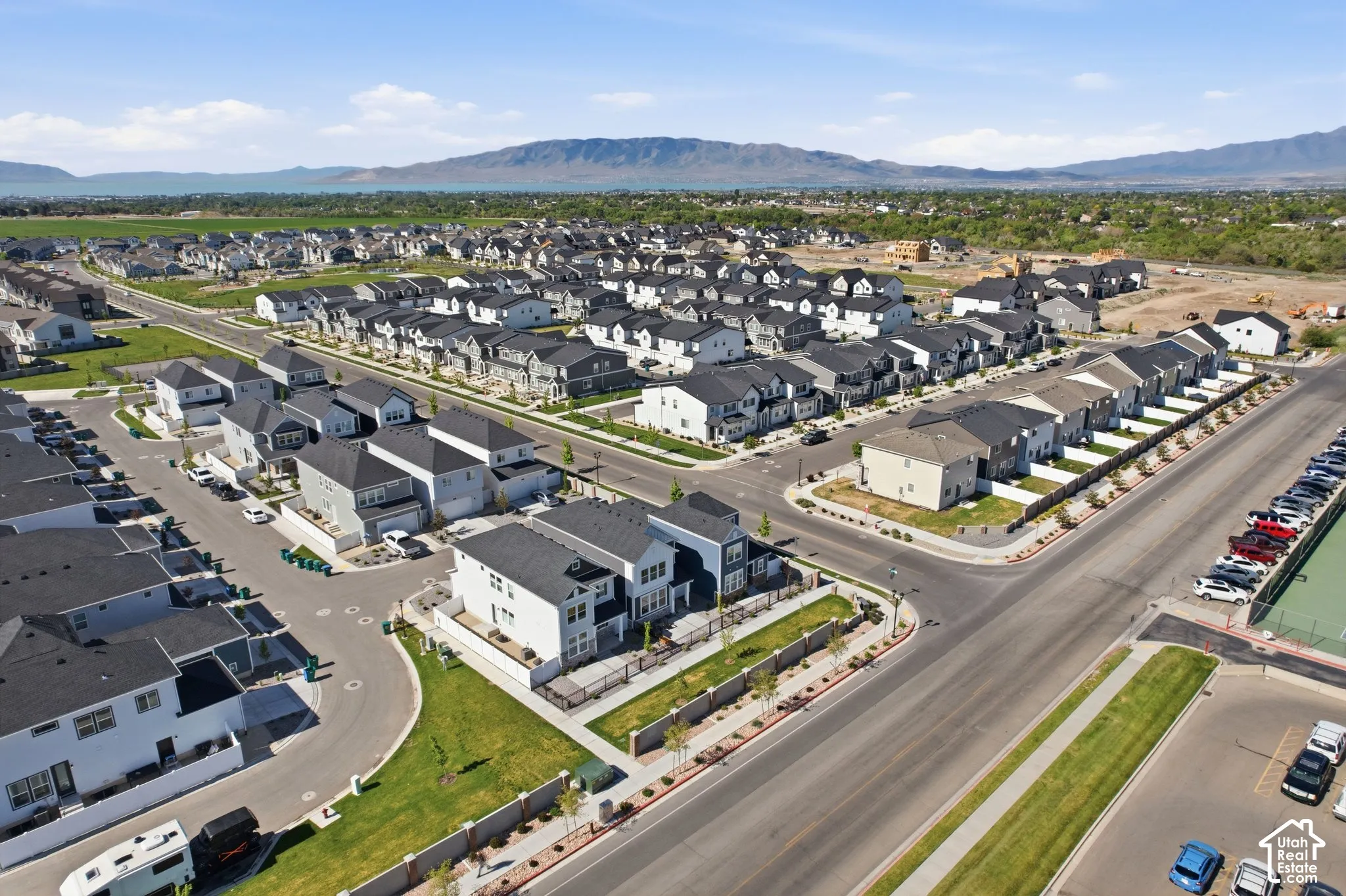 Expansive residential development featuring modern homes with dark roofs, manicured green spaces, paved streets, and mountainous terrain in the distance