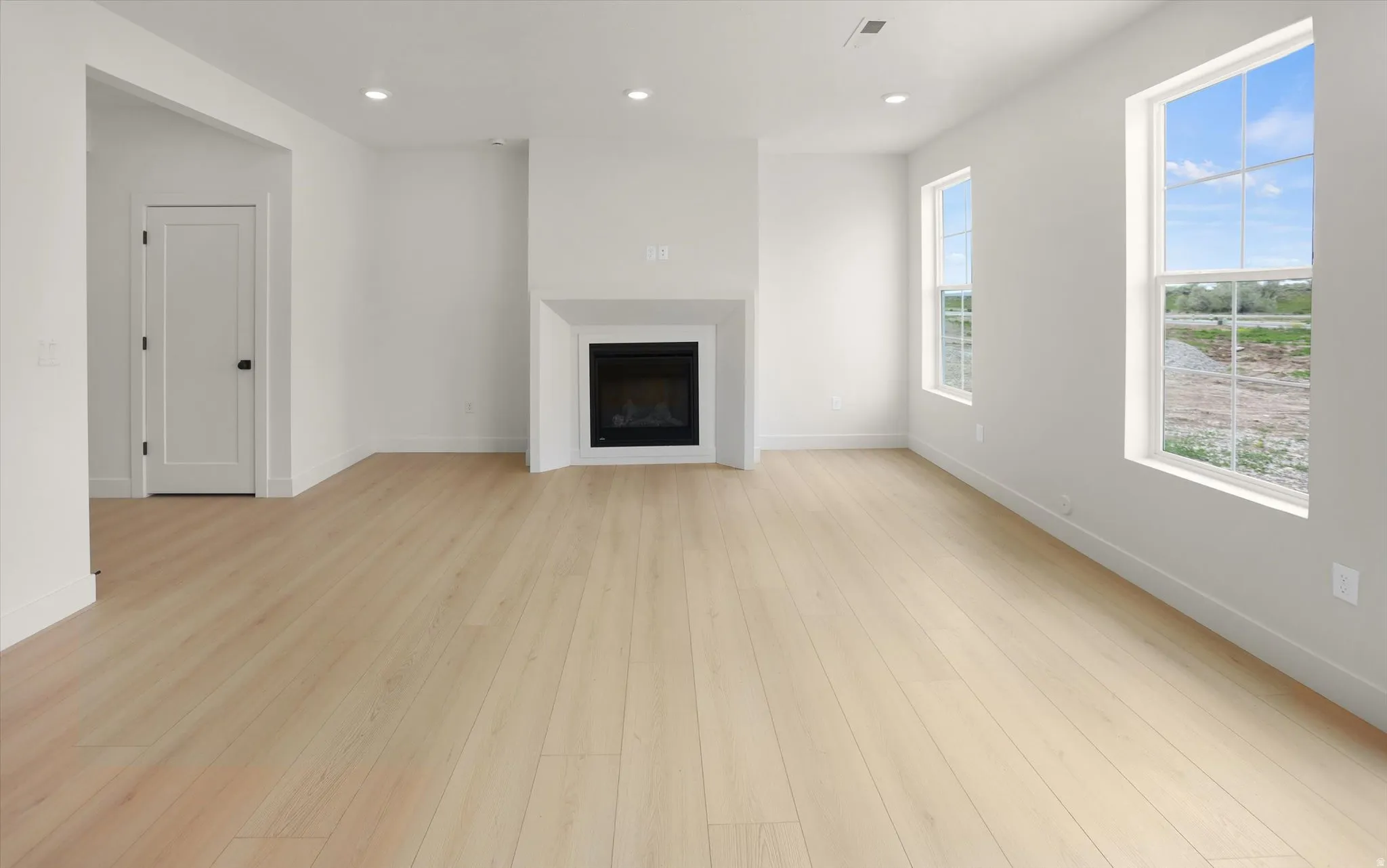 Bright living space featuring light wood-finish flooring, a contemporary fireplace with a white mantel, recessed lighting, and multiple windows providing natural light