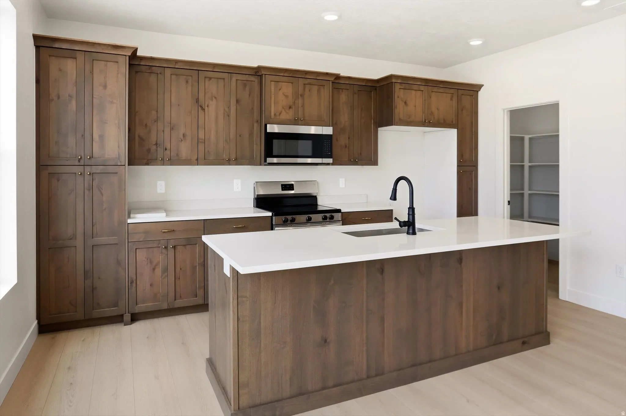 Kitchen featuring a large central island with a sink and matte black faucet, extensive wood-finish cabinetry, light-toned countertops, and stainless steel appliances