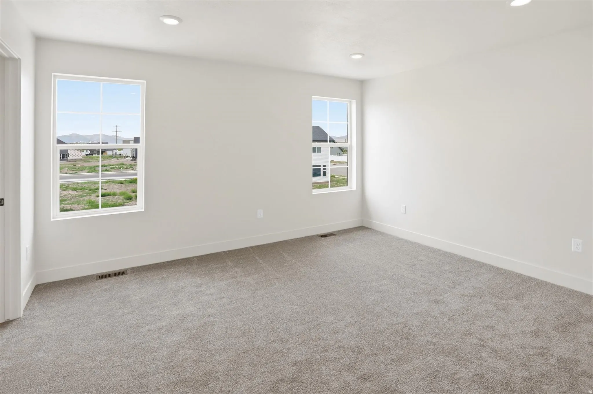 Spacious room featuring neutral carpeting, white walls, two large windows, recessed ceiling lights, and crisp white baseboards