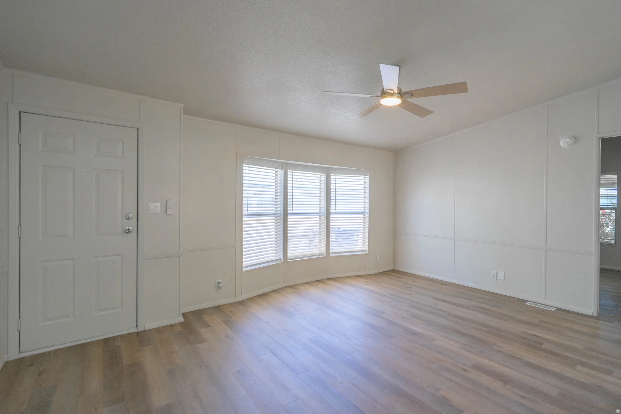 Bright living space featuring wood-finish flooring, a contemporary ceiling fan with integrated lighting, and multiple windows with horizontal blinds