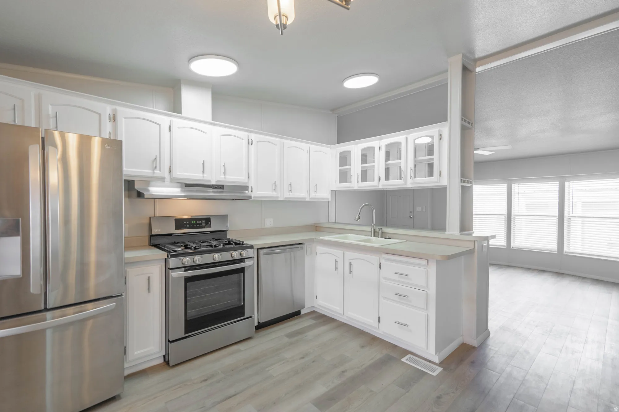 Open-concept kitchen featuring white cabinetry, stainless steel appliances, light-toned countertops, wood-finish flooring, and ample natural light from multiple windows