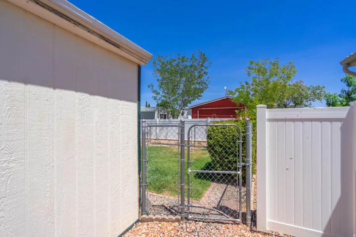 Outdoor area featuring a chain link gate opening to a grassy expanse, bordered by a white privacy fence and a building with vertical panel siding