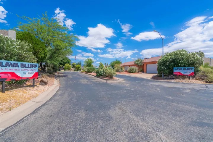 Paved entryway featuring Lava Bluff community signage, mature desert landscaping, and single-story residences with attached garages