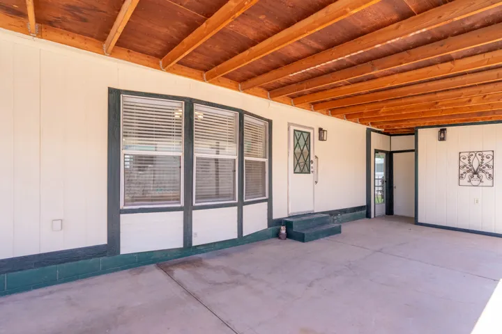 Covered concrete patio with exposed wood beam ceiling, multiple windows with blinds, a white entry door with decorative glass, and exterior wall sconces