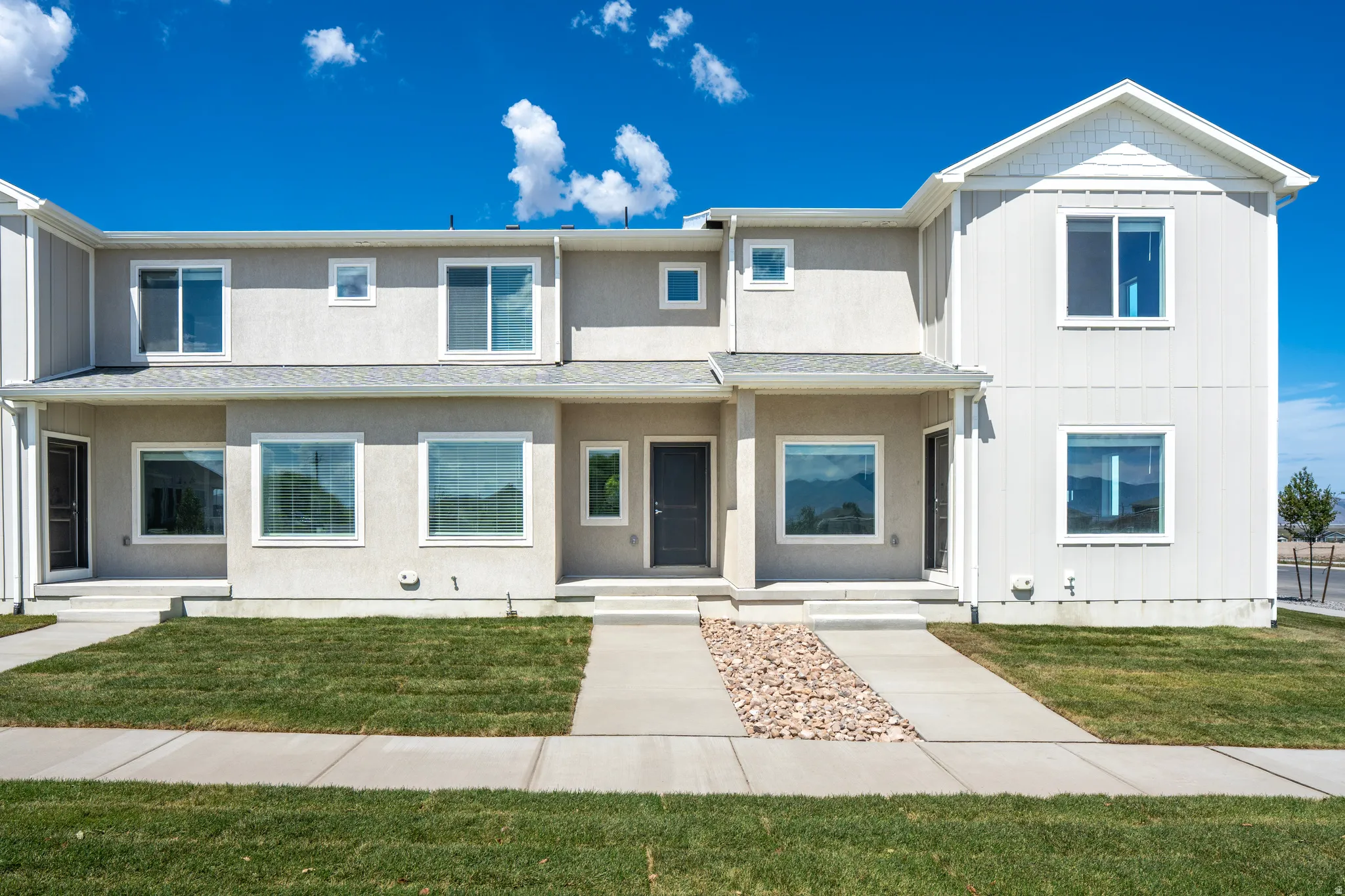 Contemporary townhouse exterior featuring a neutral stucco and siding facade, white window frames, a concrete walkway, and a manicured lawn
