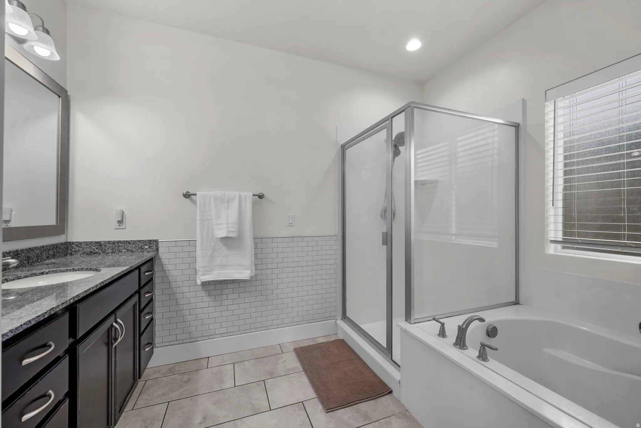 Primary bathroom featuring a glass-enclosed shower, built-in soaking tub, dark wood-finish vanity with granite countertop, and subway tile wainscoting
