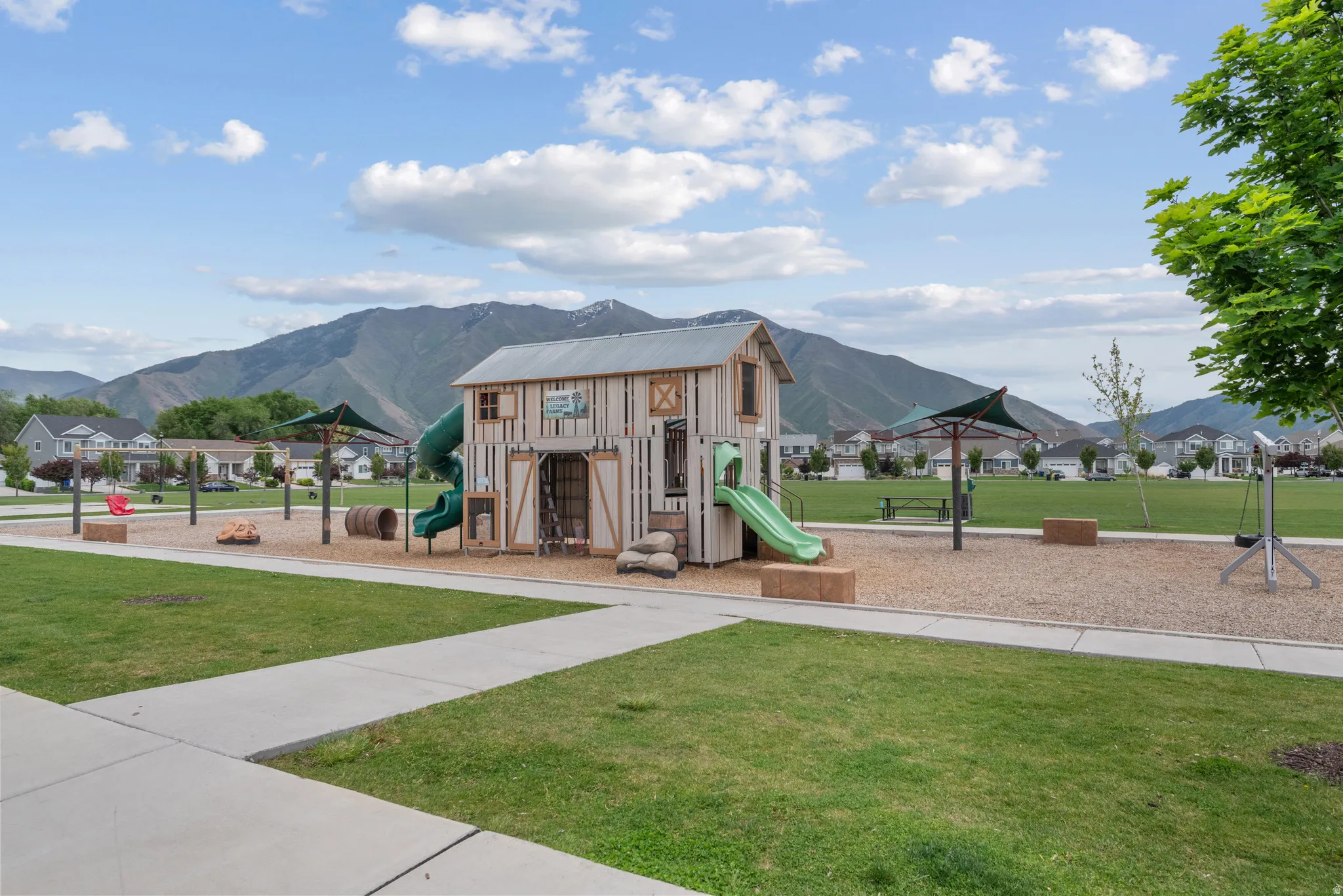 Community playground featuring a barn-style play structure with two slides, a gravel surface, swings, basketball courtm, fitness area, covered paviliion and open lawn space for picnics and soccer.