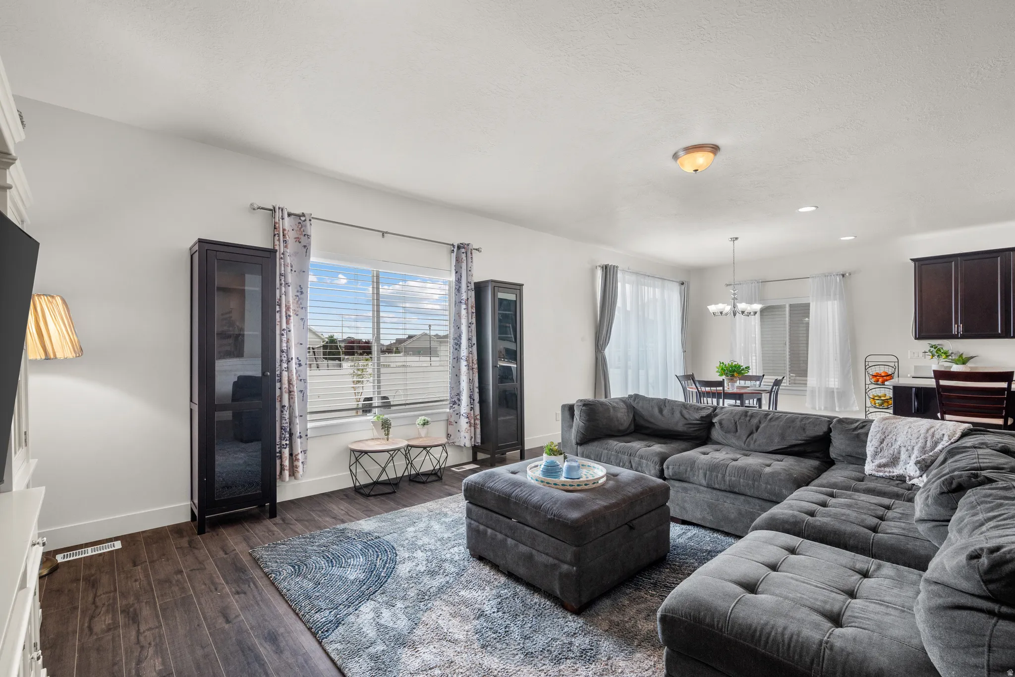 Expansive living area with dark wood-finish flooring, white walls, and a large window with blinds